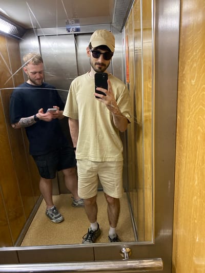 The image is a vertical, mirrored shot taken inside an elevator. The elevator's interior features polished stainless steel walls and a wooden ceiling with a distinct grain pattern. A metal handrail is visible on the right side.
Two men are visible, reflected in the mirror. The man in the foreground, wearing a black t-shirt and black cargo shorts, is holding a phone and appears to be taking the picture. He has tattoos on his left arm. His reflection is also visible.
The second man, in the upper part of the image, is wearing a light-colored t-shirt, matching shorts, a light-colored baseball cap, and sunglasses. He is also holding a phone.
The overall atmosphere is casual. The lighting suggests it's daytime, but the specific time is not evident. The location cues, such as the elevator's design, do not provide specific information about the precise location within Bielsko-Biała, Poland. No discernible text is visible in the image.