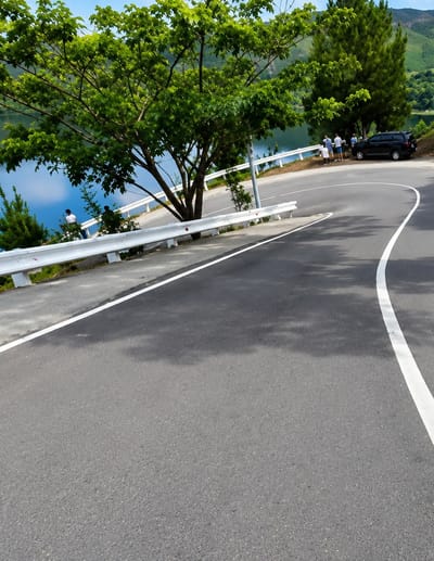 The image shows a winding paved road with a white line marking the edge. On the left side of the road, there is a body of water, likely a lake or reservoir, with a white guardrail separating it from the road. Several people are visible near the guardrail, and a dark SUV is parked on the side of the road. A large tree with green foliage is prominent in the foreground, casting shadows on the road. The background features rolling hills covered in trees, suggesting a natural or semi-rural setting. The sky is partly cloudy and light blue. This scene is located in Turpuk Sihotang, Indonesia, a location known for its natural beauty, often featuring lakes and mountainous terrain.