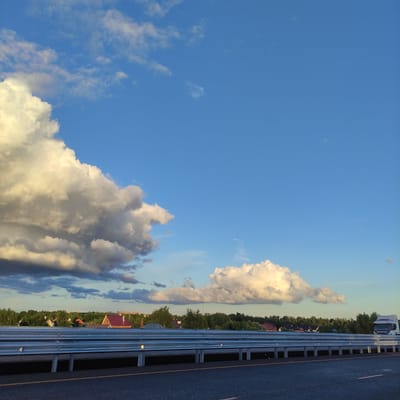 The image depicts a wide, asphalt road with a visible yellow lane marker, bordered by a silver metal guardrail. Beyond the guardrail, a line of trees forms a dense treeline, interspersed with various houses, some featuring red roofs. A large, fluffy white cloud dominates the sky on the left side of the frame, while a smaller cloud formation is visible on the right. In the distance, on the far right, a white truck is partially visible. The sky is a clear, bright blue. The overall scene suggests a view from a highway or main road passing through a semi-rural or suburban area, potentially on the outskirts of Marusino, Russia, given the context.