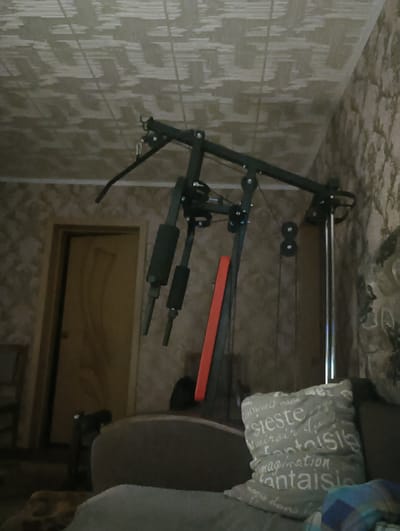 This media file shows an indoor scene, likely a living room in a residence located in Tayzhina, Russia. The primary object in focus is a home gym apparatus, a multi-station weight machine, positioned centrally. It is constructed of dark metal with various components including pulleys, cables, and padded grips. An orange element is visible on the side of the machine. To the left, a closed wooden door is partially visible, suggesting this is a room within a house. In the foreground, a portion of a sofa is visible, with a light grey decorative pillow placed on it. The pillow has white text printed on it, which appears to be in French, with words like "sieste", "fantaisie", and "imagination" discernible. The walls are covered in wallpaper with a textured, patterned design. The ceiling also has a patterned texture. The lighting suggests it is likely daytime, but the indoor setting makes it difficult to determine the exact time or weather conditions. The scene appears calm and domestic, with no people visible.
