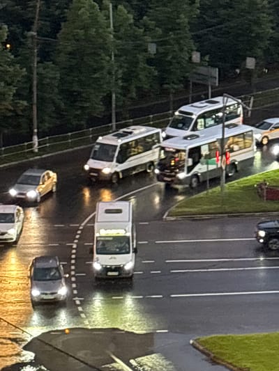 The image depicts a street scene in the city of Lesnye Polyany, Russia, during what appears to be dusk or overcast conditions, given the artificial illumination from vehicles and the dim natural light. The road surface is wet, suggesting recent rain, and reflects the headlights and streetlights.
Several vehicles are present on the road. In the foreground, a white van with its headlights illuminated is driving on the right side of the road. To the left of this van, a gray sedan is also visible with its headlights on. Further up the road, a white bus with tinted windows is driving on the left lane. Behind this bus, another white vehicle, possibly another bus or a large van, is traveling in the same direction. To the far right, a small section of a dark-colored SUV is visible. In the background, a line of trees forms a dark backdrop to the scene. The presence of streetlights and illuminated windows on the vehicles indicates that it is not fully dark yet. There are no visible pedestrians. The scene suggests a typical urban traffic situation with vehicles moving along a multi-lane road.