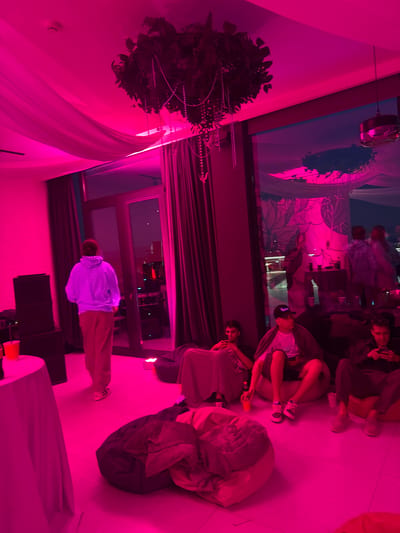The image depicts an indoor party scene illuminated by vibrant pink lighting. In the foreground, several young men are seated on beanbag chairs. One man on the left is wearing a black baseball cap and white t-shirt, looking at his phone. The man next to him is wearing a gray hoodie and also looking at his phone. To the right, another man is wearing a black jacket and sunglasses, also looking at his phone. A red plastic cup is visible near the leftmost man. In the background, a single person in a light-colored hoodie and tan pants stands near a doorway. The room has large windows offering a view of an outdoor area with some lights visible, and decorative floral elements hang from the ceiling. The overall ambiance suggests a casual social gathering. There is no indication of the specific location being in Mukhatskaro, Georgia.