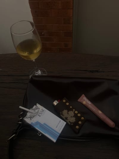 The image displays a still life scene set on a dark, rustic wooden surface, likely an outdoor table or decking, against a background featuring a segment of a red brick wall on the left and a plain light-colored wall or door on the right. A small green plant grows at the base of the brick wall. The overall lighting is dim.

In the upper left quadrant, a clear stemmed wine glass, partially filled with a pale yellow liquid consistent with white wine, stands on the wooden surface. Directly below and to the right of the glass, a dark, possibly black or deep brown, shiny material bag, resembling a clutch or small handbag with a visible zipper closure, occupies the lower portion of the frame. Arranged on top of the bag, from left to right, are three items: an open pack of "ESSE LIGHTS" brand cigarettes, white with blue details, revealing at least one cigarette; a lighter encased in a dark brown cover featuring gold-colored studs and a lighter, possibly embroidered, pattern; and a pink-toned tube labeled "rhode," likely a cosmetic product. No individuals are present. The scene is located in Thirroul, Australia.