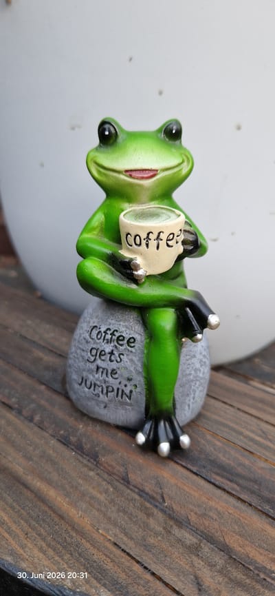 This is a close-up, low-angle, vertical shot of a small, decorative green frog figurine. The frog is depicted lying on its back on a gray, rock-like base, holding a small, light-colored mug that says "coffee" in black lettering. The frog's eyes are black and beady, and its mouth is open in a wide, smiling grin. Its limbs are black with silver tips on its fingers and toes. The base it's on has text carved into it that reads "coffee gets me jumpin'".
The frog figurine is placed on a surface of dark, weathered wooden planks. To the left of the figurine, a large, smooth, white object, possibly a pot or bowl, occupies the background. The scene appears to be outdoors, given the natural lighting. The image has a timestamp in the lower right corner: "30. Juni 2026 20:31". The text and the overall composition suggest a lighthearted and humorous mood, likely related to morning routines or the energy boost from coffee. The location context provided is Hummeltal, Germany, though no specific visual cues within the image confirm this. The lighting suggests it is late evening, close to dusk.