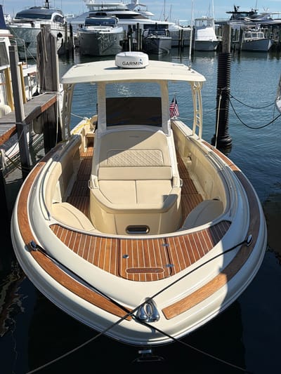 A white Chris-Craft boat is docked in the Village of Sag Harbor, USA. The boat is facing forward and is viewed from a slightly elevated angle. The interior of the boat features tan upholstered seating with diamond stitching on the backrests and a wood-planked floor. A Garmin GPS unit is visible on the white canopy above the helm. A small American flag is affixed to the starboard side of the boat. The boat is tied to a black mooring post with black ropes. In the background, other boats and yachts are docked in the harbor. The water is calm and reflects the sky and surrounding boats. The scene appears to be a sunny day.