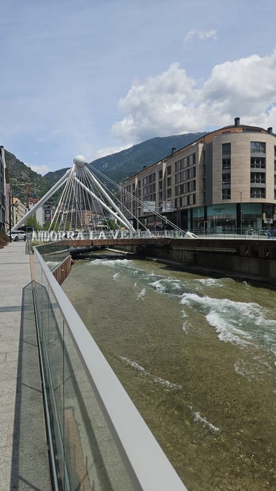 This is a daylight, eye-level shot of the Pont de Paris bridge spanning the Valira River in Andorra la Vella, Andorra. The image is taken from a paved pedestrian walkway on the near bank, looking across the river towards the city center and surrounding mountains.
In the immediate foreground, a modern railing with a light gray top rail and clear glass panels runs diagonally from the lower left to the mid-right. The walkway is paved with light-colored rectangular tiles, and shadows are cast from the railing onto the pavement on the left.
The Pont de Paris, a contemporary cable-stayed bridge, is the central feature, characterized by a prominent white spherical mast from which numerous white cables fan out to support the bridge deck. Large, white, capital letters spelling "ANDORRA LA VELLA" are mounted horizontally on the bridge structure, facing the viewer.
Beneath the bridge, the Valira River flows from left to right, exhibiting visible rapids and white foamy crests on its brownish-green surface, suggesting a shallow, rocky bed. The riverbanks are reinforced with concrete and wooden structures.
On the far side of the river, multi-story modern buildings with light-colored stone or concrete facades line the street. One building on the right features a distinctive curved corner, with large ground-floor windows displaying a "MANGO OUTLET" sign. Traffic signs are visible on poles near the buildings on the far side of the bridge.
In the mid-ground, a white SUV is partially visible on the left side of the bridge's road, and other vehicles can be seen further down the street on the right.
The background is dominated by steep, green, tree-covered mountains, rising under a bright blue sky dotted with scattered white cumulus clouds. No identifiable people are clearly visible in the scene.