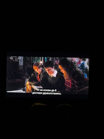 The image shows a movie screen displaying a scene with three people: Hermione Granger, Harry Potter, and Ron Weasley. They appear to be in a crowded, indoor setting. Hermione stands at the top, slightly out of focus, while Harry and Ron are positioned closer to the foreground. Harry wears glasses and a dark-colored outfit with a white collar. Ron has red hair and is dressed in a similar attire. Text is displayed on the right side of the screen, in Bulgarian, reading "И не искам да и доставя удоволствието." (I don't want to provide the pleasure.)

The environment around the screen is dark. The location is likely Suvorovo, Bulgaria, as requested. There are no other objects or actions discernible beyond those mentioned.