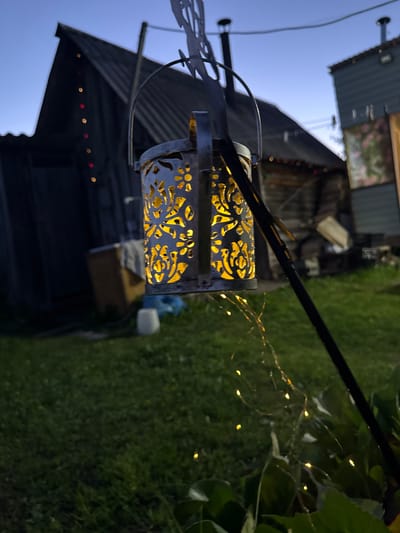 The image depicts an outdoor scene at dusk, likely in Severnyy, Russia, as suggested by its placement. In the foreground, a decorative lantern with intricate cutouts glows with warm yellow light, hanging from a dark metal pole. A string of tiny fairy lights cascades down from the pole, some of which are illuminated, adding a magical ambiance to the grass-covered yard. In the background, a rustic wooden building with a corrugated metal roof stands, adorned with a string of red and yellow lights. Adjacent to it is another building, painted green, with a window displaying a floral painting. Various objects, including a wooden crate and what appears to be blue tarp, are scattered around the yard, suggesting a lived-in, possibly storage area, or an outdoor workshop. The sky is a deep blue, transitioning from twilight to night, with a hint of clear sky visible. The overall impression is one of a tranquil evening in a rural or semi-rural setting, possibly a backyard or a small homestead.