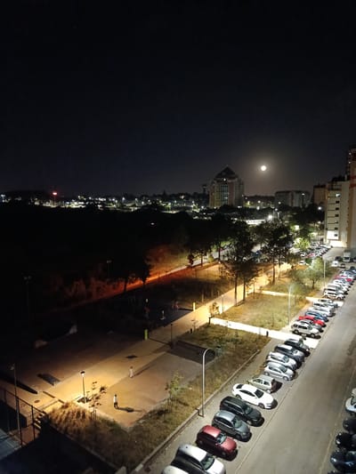 The image captures a nighttime scene in Algueirão-Mem Martins, Portugal, with a full moon prominently visible in the sky. The foreground features a parking lot filled with numerous cars of various colors and models, parked in rows alongside a street. Several streetlights illuminate the area, casting long shadows. In the middle ground, a park or recreational area is visible, with pathways, trees, and playground equipment. Two individuals can be seen walking in the park. Beyond the park, a cityscape unfolds, characterized by buildings of different heights and architectural styles. One building, a prominent, tall structure with a pyramidal top, stands out against the dark sky. The overall atmosphere is tranquil, with the urban landscape illuminated by artificial lights and the natural glow of the moon.