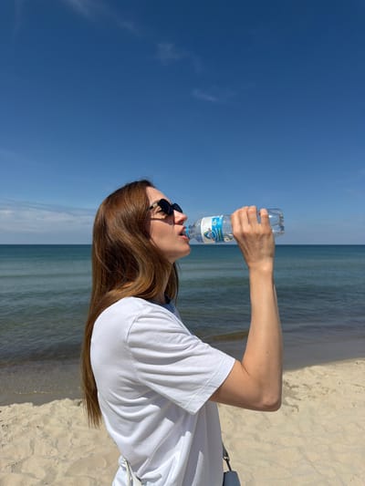 A woman with shoulder-length auburn hair and sunglasses is standing on a sandy beach and drinking from a clear plastic water bottle. She is wearing a white t-shirt. The ocean is visible behind her, with calm water and a clear blue sky overhead. The image appears to be taken during the day in a sunny climate. The location is the coast of the Baltic Sea in the village of Rybachy, Russia.