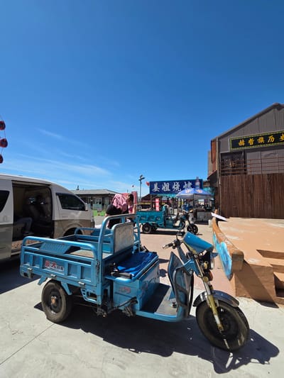 The image, taken in Jiejinkou, China, depicts a sunny daytime scene on what appears to be a street or market area. The sky is clear blue with no clouds, casting strong shadows from the objects, indicating bright sunlight, likely mid-morning or afternoon.

In the foreground and mid-ground, several light-blue electric tricycles are prominent. The closest tricycle, a three-wheeled vehicle with a small cab and an open rear cargo or passenger area, features "远大" (Yuǎndà) and "行龙" (Xínglóng) in Chinese characters, along with the English word "LADY" visible on its body. Another similar tricycle is seen further back. Partially visible on the left, a white van is parked, and a person in a pink shirt is leaning into or standing next to it. Another figure is faintly visible inside the van.

The background features buildings with dark roofs and wooden facades, suggesting shops or traditional structures. One building has a sign with "裕和铁布..." (Yùhé Tiě Bù...) written in gold characters. Blue awnings or umbrellas, possibly from street vendors or small businesses, are also visible, bearing text like "新城..." (Xīnchéng...). In the far left background, a distinctive red, circular structure, possibly part of a Ferris wheel or amusement ride, is barely visible, suggesting a recreational area nearby. The ground is a light-colored paved surface. No specific activities or events are clearly depicted, but the scene suggests daily life and commerce.