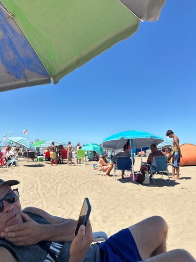 A beach scene under a clear blue sky. A row of beach umbrellas and people in chairs stretches into the distance. A person is seated in the foreground, visible from the knees up, wearing blue swim trunks. The individual is holding a cell phone. Another person's face and sunglasses are visible at the bottom left of the frame. A large green and blue beach umbrella is also visible in the foreground.