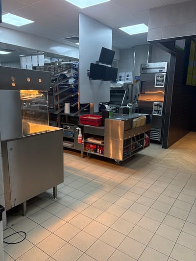 This image captures a commercial kitchen or food preparation area located in Marino Pole, Bulgaria. The scene is unoccupied, with no people present.
In the left foreground, a prominent stainless steel food holding or warming unit features a transparent front panel, illuminating its interior with a warm orange glow from overhead heating elements. The upper section of this unit displays several circular indicators.
Extending into the background, a complex arrangement of industrial stainless steel shelving units is visible, some incorporating blue accents, suggesting a storage or preparation area. Immediately to the left of the central white pillar, an additional stainless steel counter or prep area is visible.
A robust white pillar stands centrally, supporting three dark-screened monitors vertically mounted, likely serving as kitchen display systems for orders or tasks.
To the right of the central pillar, a "True" branded stainless steel commercial refrigerator is positioned. It features a digital display showing '2' and a small windowed section on its lower door. Adjacent to the refrigerator, a wall section covered in dark, rectangular tiles runs vertically, contrasting with the light-colored, smooth walls elsewhere.
In the mid-ground right, a mobile stainless steel cart equipped with multiple shelves is observed. These shelves hold various packaged items, paperwork, and what appears to be a stack of red plastic trays. A white paper towel roll and a white spray bottle with a red nozzle are also visible on nearby surfaces.
The floor in the main kitchen area consists of light-colored, square ceramic tiles with visible grout lines. Towards the far right, the flooring transitions to a wood-patterned laminate or tile. The ceiling is a drop ceiling system with evenly spaced square fluorescent light panels providing overhead illumination to the entire space. The environment appears clean, organized, and operationally ready.