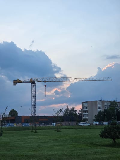 The image shows a construction site in Širvintos, Lithuania. A yellow tower crane dominates the center of the frame, with its arm extended horizontally. Behind the crane, a multi-story building with a light-colored facade and numerous windows is visible. The sky is filled with dramatic, dark clouds, tinged with pink and orange hues from what appears to be a sunset. In the foreground, a well-maintained green lawn is visible, with some small trees sparsely planted. Signs with the word "HILTUS" are present in the background, indicating a commercial or industrial area under development. A white SUV and other vehicles are parked on the street near the construction site. The overall scene suggests ongoing construction activity in an urban environment during twilight.