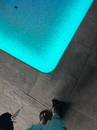 The image shows a top-down view of a swimming pool and its surrounding tiled area. The pool water is illuminated with a bright turquoise light, revealing a mosaic tile pattern on the pool floor. A dark, circular object, likely a drain or skimmer, is visible in the upper left quadrant of the pool. The pool edge is bordered by dark gray, textured tiles that appear to be laid in large rectangular slabs. In the lower right of the frame, a person's feet wearing dark flip-flops are visible, with painted toenails. The person is wearing light blue pants. A shadow cast by the person and possibly another object or person extends across the tiled area. The scene appears to be outdoors, given the natural lighting and the presence of a swimming pool. There is no specific indication of the city of Urbanización Encinares De Alcolea, Spain, within the visual elements of the image itself.