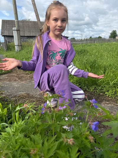 A young girl with blonde hair, wearing a purple hoodie and pants, and a pink t-shirt with a palm tree graphic, is sitting on the ground in an outdoor setting. She is looking towards the camera with a neutral expression. Her right arm is raised, palm facing outwards, as if waving or posing. She is surrounded by lush green grass and wildflowers. In the background, there is a wooden fence and a rustic building, suggesting a rural or village environment. The sky is overcast with clouds. The overall mood is relaxed and casual, possibly during a daytime outdoor activity. Text on the t-shirt is partially visible and appears to say "SEA SURF". The location is identified as Большой Турыш, Russia.