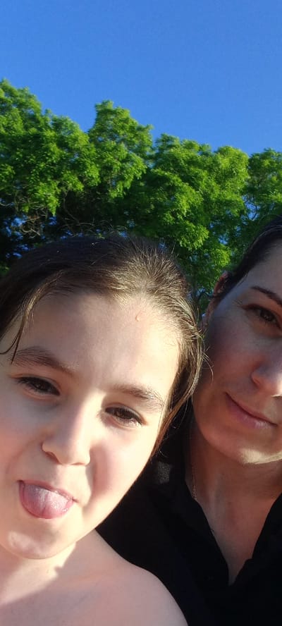 This is a close-up, selfie-style photograph of two people with bright green trees and a blue sky in the background. The person on the left, a younger individual, has dark hair and is sticking out their tongue, with a small droplet of water visible on their forehead. The person on the right, an adult, has dark hair and is smiling, with a delicate gold chain visible on their neck. Both individuals appear to be outdoors. The location of Gwandra, Abkhazia is not visible or inferable from the image.