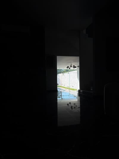 The image depicts a dimly lit interior space in Villahermosa, Mexico, looking through a rectangular opening towards a brightly illuminated exterior. The interior floor is composed of highly reflective dark, possibly black, tiles that show reflections of the outdoor light. Above the opening, a modern chandelier with multiple dark, spherical light fixtures is visible. To the left of the opening, a dark-framed picture or mirror hangs on a lighter wall section. To the right, a staircase with visible steps ascends from the floor level.
The exterior scene features a light blue swimming pool equipped with a two-rung ladder for access. Behind the pool, a textured white or light-colored wall with vertical patterns forms a barrier, beyond which green foliage, likely trees or bushes, is discernible under bright daylight conditions. There are no individuals, overt actions, or interactions present within the frame.
