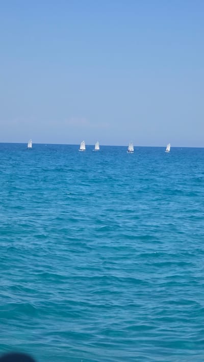This is a rotated aerial photograph showing a line of small sailboats with white sails on a bright blue sea. The sea has a textured surface with visible waves. The sky is a clear, pale blue, meeting the sea at a distinct horizon line that runs vertically down the center of the image due to its rotation. There are at least six sailboats visible, spaced out along the horizon. The boats are all facing in the same direction, suggesting they are part of a race or a coordinated sailing activity. The overall impression is one of a peaceful, sunny day at sea, likely during daylight hours. There is no visible text in the image.