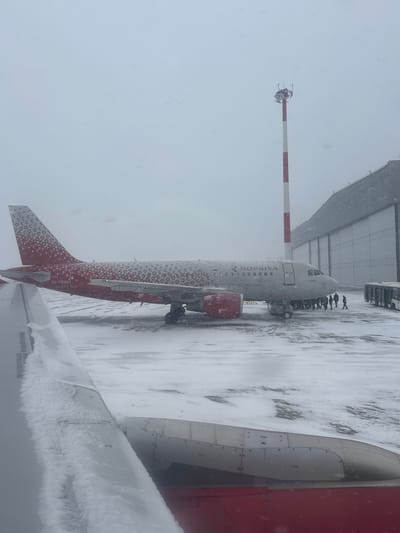 The image captures an airport apron at Емельяново (аэропорт), Russia, during an ongoing snowfall. A Rossiya Airlines Airbus aircraft, registration RA-73190, is stationary on the snow-covered tarmac. The aircraft features a light gray fuselage with a distinctive red and white patterned livery on its tail and engine nacelles, and "ROSSIYA" lettering on its side. Approximately 10 to 15 individuals, dressed in dark winter clothing, are congregated near the front right main cabin door of the aircraft, with some appearing to move towards the right side of the frame. To the right of the aircraft, two white airport buses are partially visible. In the background, a tall red and white striped mast or communication tower stands adjacent to a large, light-colored building, likely a hangar or terminal, which extends across the right side of the background. The foreground reveals a snow-covered aircraft wing and a portion of a red engine cowling, indicating the image was taken from another parked aircraft. The entire scene is under overcast skies, with visible snowflakes falling and accumulated snow covering all exposed surfaces.
