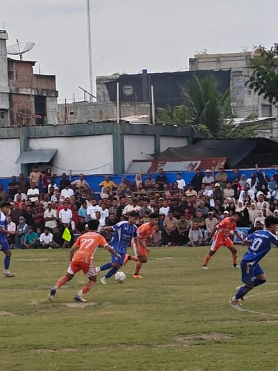 This image captures a football match in progress on a grassy field, likely in Desa Meunasah Blang, Indonesia. The foreground shows several players in action. Two players in orange jerseys are prominent, one with the number 77 on his back and the other in motion. A player in a blue jersey with the number 10 is also visible, running on the field. A football is present near the players. In the background, a large crowd of spectators is gathered, seated and standing behind a blue barrier. The spectators are diverse in age and gender, many dressed in casual attire, with some women wearing headscarves. Behind the spectators, a cityscape is visible, characterized by buildings with varying architectural styles, including brick structures and concrete buildings. Some structures appear weathered, and there are various antennas and satellite dishes on rooftops, indicative of urban or semi-urban infrastructure. The sky is overcast, suggesting diffused lighting conditions. The overall scene depicts a community sporting event with active participation and an engaged audience in an Indonesian setting.