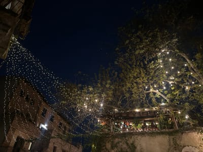 This image depicts an outdoor night scene in Santa Cruz Aquiahuac, Mexico. A dark blue night sky is visible in the upper half of the frame. On the left, a multi-story building constructed of light-colored masonry, possibly brick or stone, features multiple dark, rectangular window openings. From an upper architectural element of this building, numerous strands of small, warm-toned string lights are suspended diagonally downwards and across the scene. On the right, large trees with discernible foliage and branches are extensively decorated with similar string lights, illuminating parts of their canopy. Below these trees, in the mid-ground, a wooden pergola or covered outdoor structure is present, also adorned with string lights. Underneath this structure, indistinct colorful objects, possibly banners or decorative elements, are visible. A low, light-colored wall with climbing vegetation runs across the lower mid-ground. The area is illuminated predominantly by the decorative string lights. No individuals, specific actions, or interactions are discernible within the frame.