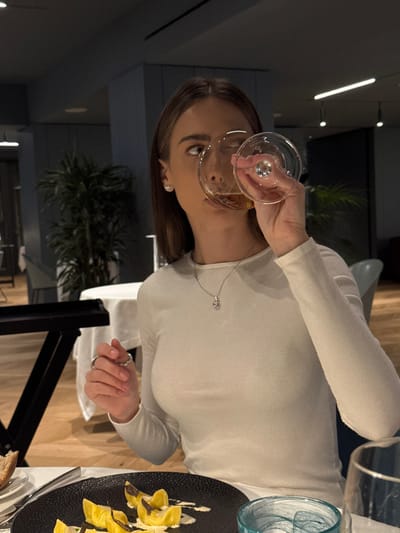 A young adult female is depicted seated at a dining table in an upscale restaurant in Torbole, Italy. She is wearing a long-sleeved white top, a silver necklace with a rectangular pendant, and a ring on her left ring finger, with stud earrings visible on both ears. Her head is tilted slightly back as she holds a clear stemmed glass containing an amber liquid to her mouth, appearing to consume the beverage. Her gaze is directed upwards and to the right. In the foreground, on a dark, textured plate, are several pieces of yellow pasta, likely ravioli or tortellini, arranged in a white sauce and garnished with dark flakes, possibly truffles. A blue-tinted glass is positioned to the right of the plate, and a small white plate with a piece of bread is to the left. The table is covered with a white tablecloth. The background features a dimly lit interior with blue-grey architectural elements, recessed lighting, and large potted green plants. Other dining tables are partially visible in the blurred distance. The flooring appears to be light-colored wood.