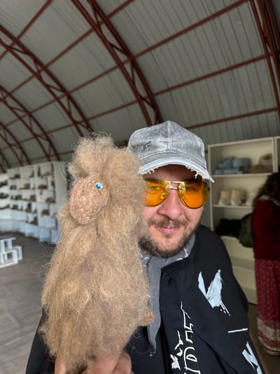 A man is holding a fluffy, light brown, handmade doll with a single blue eye. He is wearing a grey baseball cap, orange tinted sunglasses, and a dark long-sleeved shirt with a white graphic print. He is smiling and looking at the camera. In the background, there are shelves stocked with various items, possibly textiles or crafts. The ceiling structure appears to be made of metal beams and light-colored panels. The overall setting suggests an indoor market, workshop, or retail space, potentially in Tri Ozera, Russia.