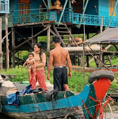 In the city of Soutr Nikom, Cambodia, a woman stands on a boat holding a baby. She is wearing a tan long-sleeved shirt and red patterned pants. A man, shirtless and wearing black shorts, stands to her right, facing away from the camera. The boat is old and weathered, painted in shades of blue and green, with a red painted bow. Various objects, including a tire and some clothing, are present in the boat. In the background, a stilted wooden structure painted blue, with red and yellow accents, rises above lush green vegetation and murky water. The architecture suggests a village built on stilts, common in areas with fluctuating water levels. The scene appears to depict a moment of daily life in this community.