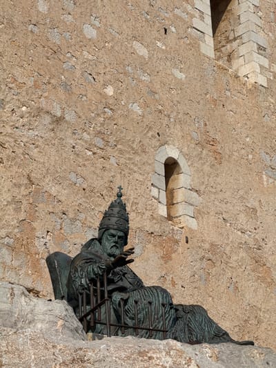 The image displays a bronze statue of a robed figure, likely a pope or a high-ranking cleric, mounted on a textured stone wall in Peníscola, Spain. The figure wears a detailed papal tiara or mitre and appears to be in a blessing or speaking pose, with one hand raised. The statue exhibits a dark, oxidized green patina, indicative of aged bronze.
The wall behind the statue is composed of light-colored, roughly textured stone or stucco, interspersed with various sizes of aggregate and lighter gray stone patches. To the upper left of the statue, an architectural detail is visible: a small, semi-circular niche or recess with a drain-like opening, framed by light-colored, chevron-patterned stones. Further to the left, another section of the wall shows similar masonry work. On the immediate right, the statue is partially framed by a natural, rugged grey rock formation, suggesting the wall is integrated into a cliff face or substantial existing rock.
The scene is illuminated by bright daylight, casting distinct shadows that highlight the sculptural forms and the uneven texture of the stone. The expression on the statue's face is solemn. Given the location of Peníscola, this statue is likely a depiction of Pope Benedict XIII, known as Papa Luna, who resided in the town's famous castle. There is no visible text in the image.