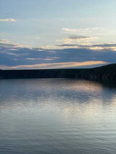 The scene features a body of water, possibly a lake, reflecting the sky. Its surface is ruffled by light waves, creating a textured appearance. A dark, vegetated landmass stretches along the water's edge. The sky transitions from a clear, light blue to areas with elongated clouds tinged with golden light, suggesting either sunrise or sunset. The overall atmosphere is calm and serene.