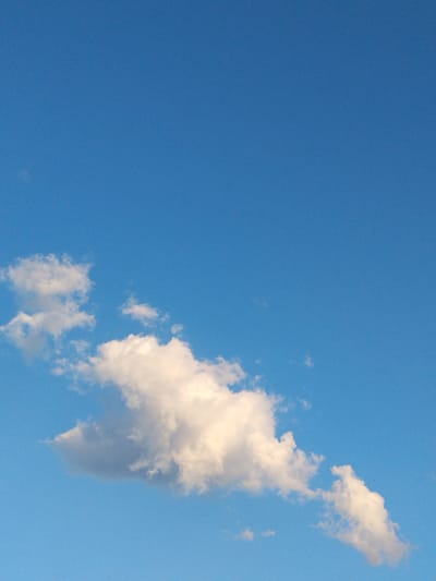 The image is a vertical, eye-level shot of a clear blue sky with scattered white clouds. A large, fluffy cloud is centered in the lower half of the frame, with smaller wisps of clouds to its left and right. The lighting suggests it is daytime. There is no visible indication of the city of Mikhailovka, Russia, or any people or specific objects within the frame. The scene is solely focused on the sky and clouds.