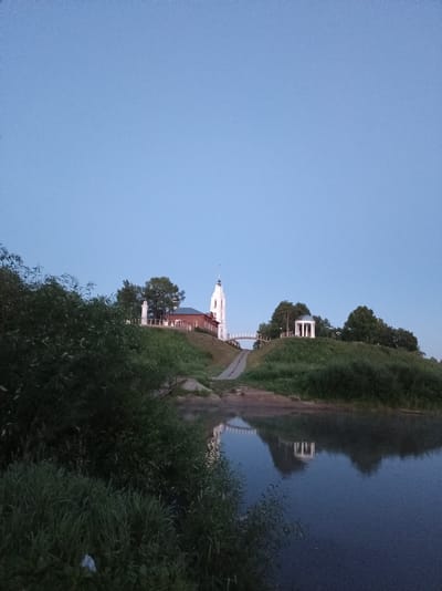 This is a photograph of a riverbank in Buy, Russia. In the background, on a hill, there is a white bell tower and a red brick building, likely a church, with a white spire. There is also a white rotunda structure on the hill, and a bridge connecting two parts of the hill. The sky is a clear, pale blue, indicating it is either early morning or late evening. The foreground is dominated by lush green foliage and grass. The river in the foreground reflects the sky and the structures on the hill. There are no people or animals visible in the image.