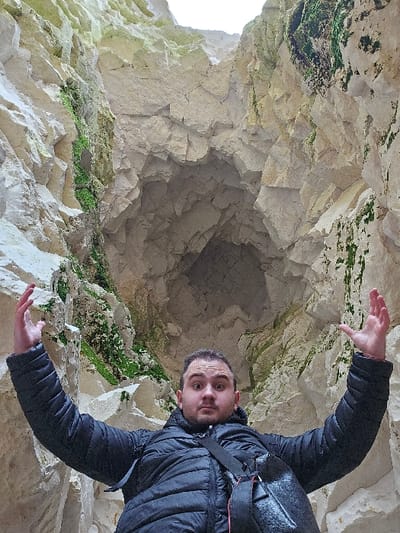 An adult male stands centrally within a deep, narrow chasm formed by towering white chalk cliffs in Thanet, United Kingdom. The man, appearing to be in his late 20s to early 40s, has short dark hair and stubble. He is wearing a black quilted hooded jacket and a dark shoulder bag diagonally across his body. His expression is wide-eyed with a slightly open mouth, and both arms are raised high, palms open and fingers splayed, creating a gesture of perceived awe or scale. The chalk cliff faces rise steeply on both sides, displaying jagged, fractured surfaces, and recede into the background. Patches of green moss and algae adhere to various sections of the light-colored rock. The sky, visible at the top of the chasm, appears bright, providing diffused overhead lighting.