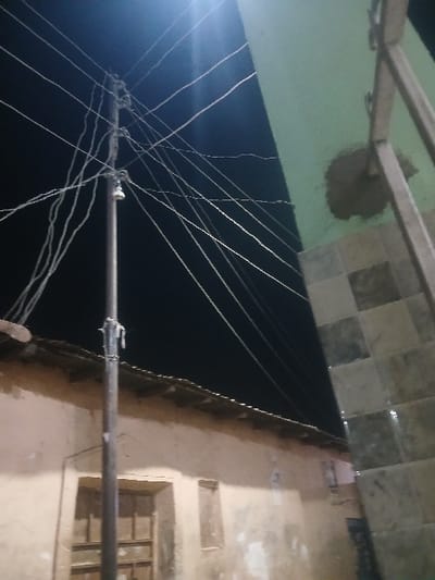 An upward shot at night shows a tangle of electrical wires connected to a horizontal pole. Adjacent to the wires is a beige building with a window. Above the wires are a green platform and a tiled overhang. The platform has a spot of repair work. The scene is mostly dark, emphasizing the wires' chaotic arrangement.