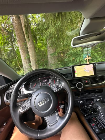 The image shows the interior of an Audi vehicle parked in a forested area. The perspective is from the driver's seat, looking towards the dashboard and windshield. Through the windshield, tall trees with green foliage are visible, suggesting an outdoor, likely rural setting.
Essential details include:
- **Vehicle Interior:** The dashboard features an infotainment screen displaying a navigation map. The steering wheel, complete with the Audi logo, is prominent. Various buttons, controls, and the gear shifter are visible. A wooden cross is hanging from the rearview mirror. A phone mount with a phone is attached to the dashboard. The interior appears to be leather and wood trim.
- **Driver's Feet:** The driver's feet are visible, one wearing a Nike sandal.
- **Exterior Environment:** The background consists of dense trees and lush green foliage. Sunlight filters through the trees, indicating daylight.
While the image provides a clear view of the car's interior and its immediate surroundings, there is no explicit visual information within the image that directly indicates the location as Oberammergau, Germany. The description of the scene is limited to what is directly observable in the photograph.