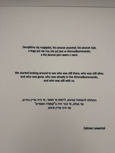 The image displays a light gray wall with black text printed on it. The text is presented in three languages: Polish, English, and Hebrew. The Polish text reads: "Zaczęliśmy się rozglądać, kto jeszcze pozostał, kto jeszcze żyje, a kogo już nie ma, kto już jest w Himmelkommando, a kto jeszcze jest razem z nami." The English text reads: "We started looking around to see who was still there, who was still alive, and who was gone, who was already in the Himmelkommando, and who was still with us." The Hebrew text reads: "התחלנו להסתכל סביבנו, לראות מי נשאר, מי היה עדין בחיים, ומי נעלם, מי כבר היה ב"קומנדו השמייים" ומי היה עדיין איתנו." Below the Hebrew text, the name "Zalmen Lewental" is printed. The image does not contain any information or visual cues to indicate a specific location within the city of Oświęcim, Poland. The scene is static and consists only of the text on the wall.