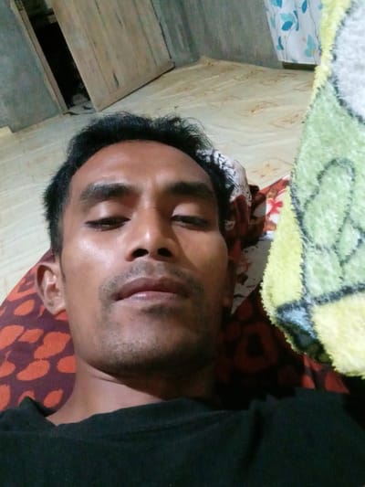 The image shows a close-up, slightly distorted view of a person's face as they lie down. The person is looking upwards and to the side, with their mouth slightly open. They have dark hair and what appears to be stubble on their chin and upper lip. Their eyes are partially closed, suggesting they might be tired or sleepy.
The person is lying on a bed or similar surface, with patterned fabric visible beneath their head, featuring an abstract design in shades of orange and dark purple. A green and yellow patterned blanket is draped over the lower portion of the frame. The background shows a light-colored wooden surface, possibly a wall or furniture, and a darker opening, which could be a small closet or shelf.
The lighting appears to be artificial, casting a warm glow on the person's face. The overall impression is one of a casual, intimate moment. There is no visible text in the image, and no clear indicators of the time of day or specific weather conditions. The setting is implied to be indoors, likely a bedroom, given the bedding. The person's expression could be interpreted as relaxed or contemplative. The location context provided is Makmur, Indonesia.