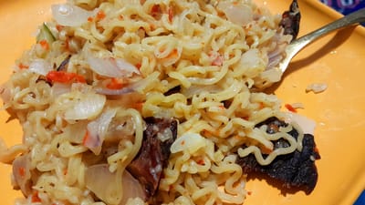 This close-up image shows a plate of noodles, likely instant ramen, served on a bright orange or deep yellow plate. The noodles are curly, pale yellow, and appear to be coated in a light-colored sauce. Mixed throughout the noodles are various ingredients, including numerous slices of translucent white and light purple onions, and small, bright red pieces that resemble chopped chili peppers or bell peppers. Several dark, irregularly shaped pieces, possibly meat or another cooked ingredient, are also visible within the dish and on the plate. A small piece of green vegetable, possibly cucumber, can be seen on the left side.
A silver-colored fork with a dark handle is partially visible on the right side, resting in the food, indicating the meal is ready to be eaten or is in progress. The lighting suggests an indoor setting. There is no visible text in the image. The context provided indicates the location is Umuigbekwo, Nigeria.