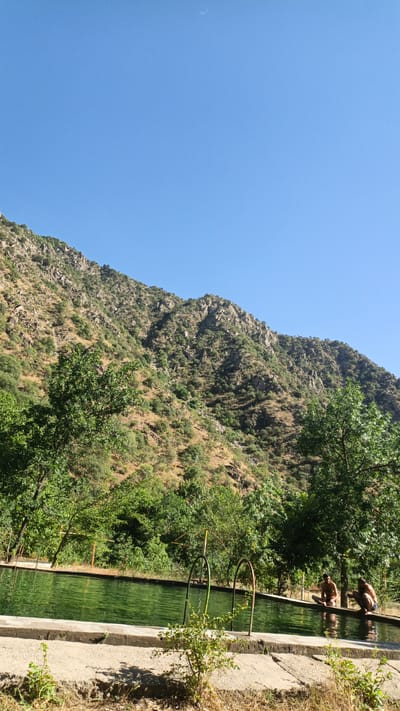 In a mountain landscape near Mirabad, Tajikistan, a rectangular, shallow pool with green water is bordered by a concrete edge. Two men, wearing swim trunks, are seated on the edge of the pool with their feet submerged in the water. Lush green trees surround the pool and extend up the arid, rocky mountainside that dominates the background. The sky above is a clear, bright blue.