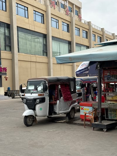 The image depicts a street scene in Bavet Quarter, Cambodia, featuring a white tuk-tuk as the primary subject, parked alongside a building with a beige facade and numerous windows. Other tuk-tuks are visible in the background. A blue awning bearing the word "VIDA" is partially visible above the vehicle. Street signs in Khmer are displayed. The tuk-tuk has an LPG sticker on the front. The overall scene suggests a typical urban environment in Southeast Asia.