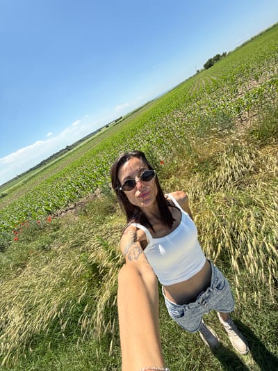 A young adult female is depicted taking a selfie in an agricultural field located in Slavovitsa, Bulgaria, under a clear, bright blue sky with minimal white clouds. The image is captured from a wide-angle, slightly high perspective, with a noticeable diagonal tilt to the right. The subject has dark hair and wears dark, hexagonal sunglasses, a white sleeveless crop top, light-wash denim shorts, and light-colored sneakers with white socks. Visible tattoos include a sun/moon crescent design on her left shoulder and intricate patterns on both forearms. She has a philtrum piercing and is shown with a slight pouting expression.
The immediate foreground consists of tall, straw-colored grasses with seed heads, interspersed with green ground cover. Beyond this, a large field of uniformly planted young green crops, likely corn, extends in discernible rows towards the horizon. To the mid-left of the frame, a cluster of red poppy flowers is visible within the vegetation. The distant background reveals gently rolling terrain with more green fields and trees. Strong daylight illuminates the scene, creating defined shadows.