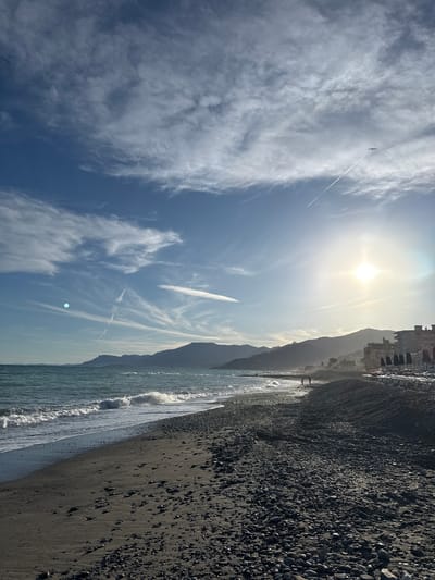 This is a wide-angle, vertical shot of a beach in Vallecrosia al Mare, Italy, with the Ligurian Sea on the left and buildings and mountains on the right. The sky is a vibrant blue with wispy white clouds and a bright sun creating a lens flare. A plane's contrail stretches across the sky, and a bird is silhouetted against it. The sea has small waves breaking on the shore. The beach itself is composed of dark sand and pebbles, with a line of beach chairs and umbrellas set up near the buildings. Two people are visible walking on the beach in the distance, and another person appears to be standing near the water. The background features rolling hills or mountains, some of which are covered in vegetation. The scene is bathed in sunlight, suggesting it is daytime.