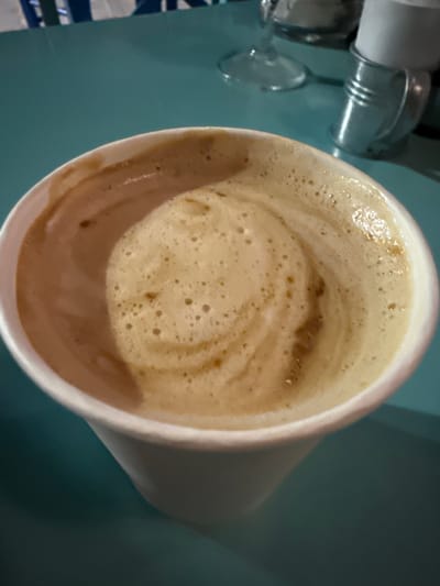 A close-up, overhead shot shows a white paper cup filled with a frothy coffee beverage. The coffee has a creamy, light brown foam with swirls of a darker brown, indicating it may be a latte or cappuccino. The cup is positioned on a teal-colored surface, likely a table. In the background, out of focus, are elements that suggest an outdoor or casual dining setting. These include a glimpse of a striped fabric in blue and white, a wine glass, and a small metal container holding white paper or napkins. The lighting suggests it is daytime, possibly outdoors given the reflections and color palette. The scene is devoid of any people or specific identifiable landmarks of Medinski Krš, Montenegro, beyond what can be inferred from the general setting.