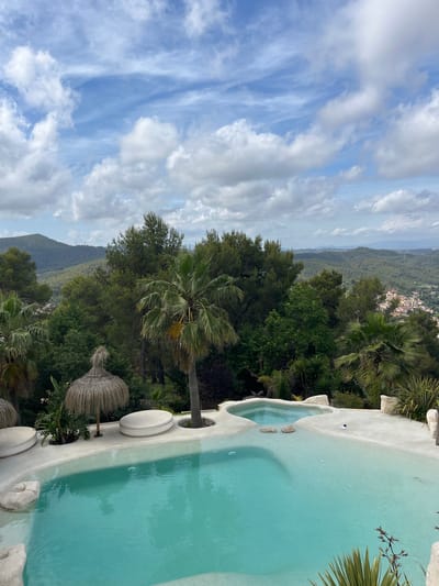 The image shows a view from a terrace overlooking a lush green landscape with a town in the distance. In the foreground, there is a swimming pool with turquoise water and a surrounding white deck. Several white, rounded lounge chairs and a thatched umbrella are placed on the deck. Palm trees and other greenery are visible around the pool area. In the background, rolling hills covered with trees descend towards a town with red-tiled roofs. The sky is partly cloudy with scattered white clouds against a blue background. The location is identified as Can Güell, Spain. There are no people or animals visible in the image. The scene depicts a tranquil, scenic outdoor environment with a focus on a luxurious swimming pool area.