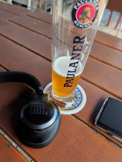 An outdoor wooden table features a tall glass of Paulaner beer, a pair of black JBL headphones, and a small, black, textured object, possibly a phone or a wallet. The beer glass, which has a foamy head, rests on a Paulaner coaster. The headphones are positioned to the left of the glass. The scene appears to be a casual setting, perhaps a beer garden or outdoor cafe. The overall lighting suggests daytime. The image does not depict any people or specific actions. The location is identified as Denkendorf, Germany.