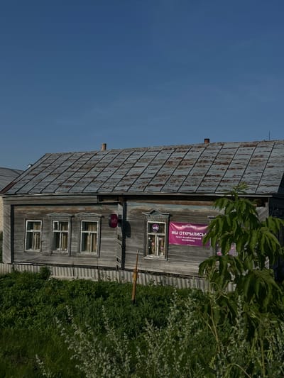 The image depicts a weathered, wooden, two-story building with a corrugated metal roof. The building is situated in an outdoor environment with a clear blue sky overhead and lush green vegetation in the foreground. Several windows are visible on the building's facade, some adorned with decorative trim. A pink banner with Russian text and the "wb" logo is prominently displayed on the right side of the building. There are no people visible in the image. The overall impression is one of rural or small-town architecture, possibly in a setting such as the city of Mazan, Russia, given the context.