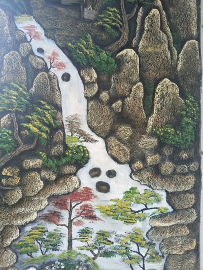 This is a textured, three-dimensional artwork depicting a stylized mountainous landscape with a winding river or waterfall. The scene is presented in a bas-relief or highly textured painting style, giving it a tactile quality.
The central feature is a white river or waterfall that flows from the top, meandering downwards through the composition in an S-curve. Several dark, circular or oval shapes are visible within the white water, possibly representing deep pools or rocks submerged in the current.
On either side of the waterway, rugged and textured rock formations rise, colored predominantly in shades of brown, grey, and ochre, resembling craggy mountains and large boulders. These rocks create a sense of depth and scale within the scene.
Interspersed among the rock formations and along the riverbanks are various trees and patches of foliage. Trees with vibrant green, golden yellow, and deep red leaves are strategically placed throughout the landscape. The red-leafed trees, in particular, stand out, suggesting either autumn colors or specific species like maples. Smaller green shrubs and areas with tiny yellow flowers add detail near the water's edge.
The artwork evokes a sense of serene natural beauty, reminiscent of traditional East Asian landscape art. There are no people, specific activities, or discernible text present in the image. The lighting suggests a clear daytime setting. The artwork is located in Bangsri, Indonesia.