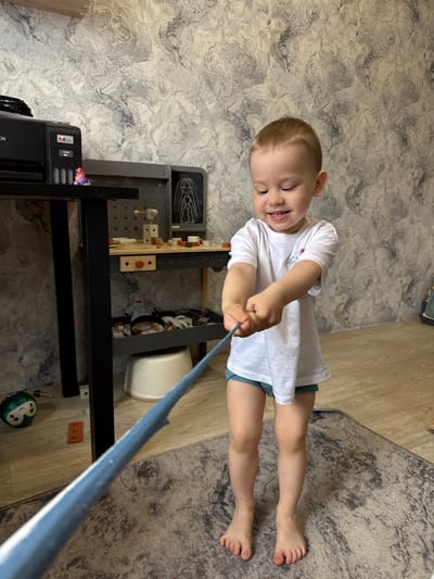A young child with short, light-brown hair, wearing a white t-shirt and blue shorts, is pulling on a blue-colored pole. The child is positioned on the floor, which is covered partly by a gray rug and partly by light-colored hardwood flooring. The floor also features wallpaper with a marbled pattern. In the background, there's a black shelving unit and a white bucket or container. The exact location in the city of Aristovo, Russia, is not discernable from the image.