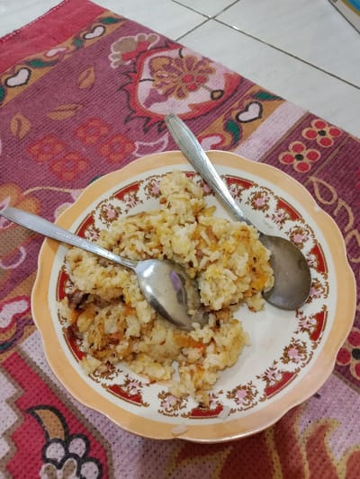A plate of fried rice, consistent with Nasi Goreng, is positioned centrally on a patterned maroon-colored rug. The rice is light brown and white, mixed with discernible pieces of fried shallots or onions, and possibly other small ingredients. Two silver-colored metal spoons rest on the rice. The spoon on the left displays "Bon appétit" engraved on its handle. The plate has a scalloped edge, an orange rim, and a white interior decorated with a repeating floral pattern featuring red and gold flowers with green accents. A small section of white tiled flooring is visible in the upper right background, indicating an indoor setting. The scene is located in Brangsong, Indonesia.